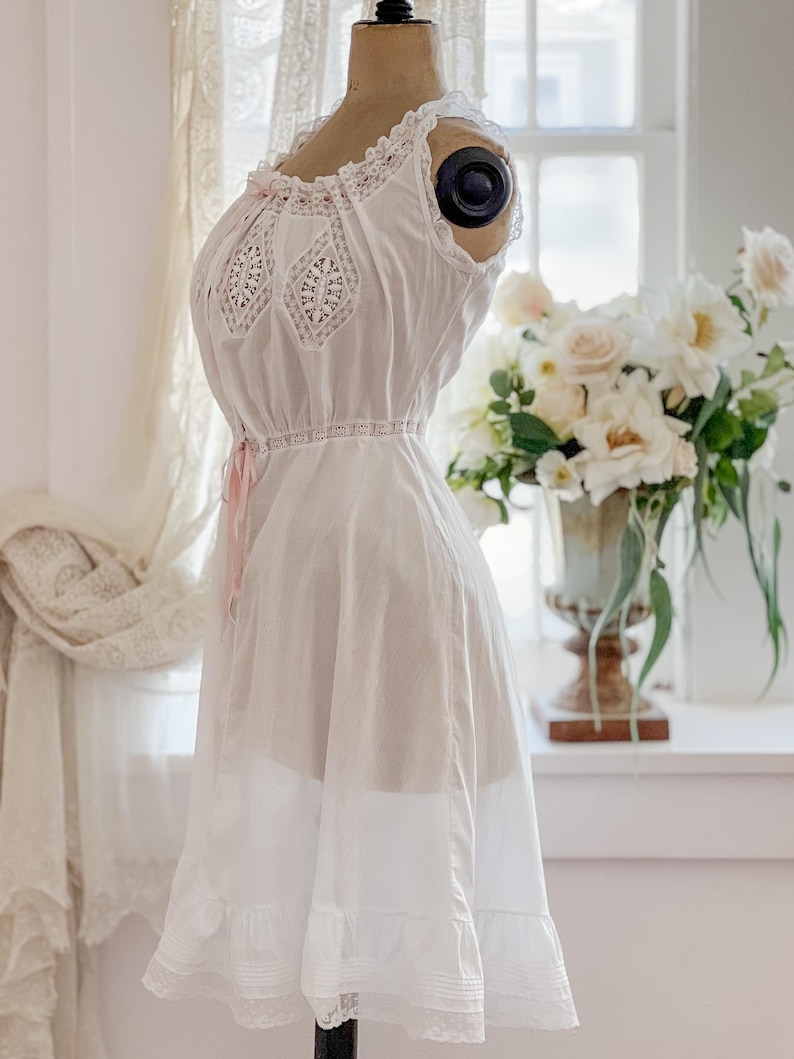 May include: A white vintage-style nightgown on a mannequin. The sleeveless gown features lace detailing on the bodice and hem, with a pink ribbon accent at the waist. The dress is displayed in front of a window with a floral arrangement.