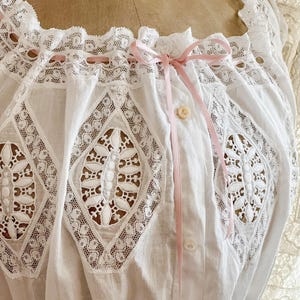 May include: Close-up of a white vintage blouse with intricate lace detailing. The blouse features a square neckline, diamond-shaped lace inserts, and a delicate pink ribbon bow. A single button and loop closure is visible.