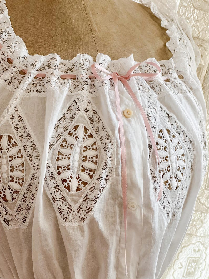 May include: Close-up of a white vintage blouse with intricate lace detailing. The blouse features a square neckline, diamond-shaped lace inserts, and a delicate pink ribbon bow. A single button and loop closure is visible.