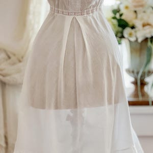 May include: A white vintage dress with delicate lace and pleating details. The dress features a high neckline with vertical stitching and a pink lace band at the waist. The skirt has a layered design with a ruffled hem, creating a flowing silhouette.