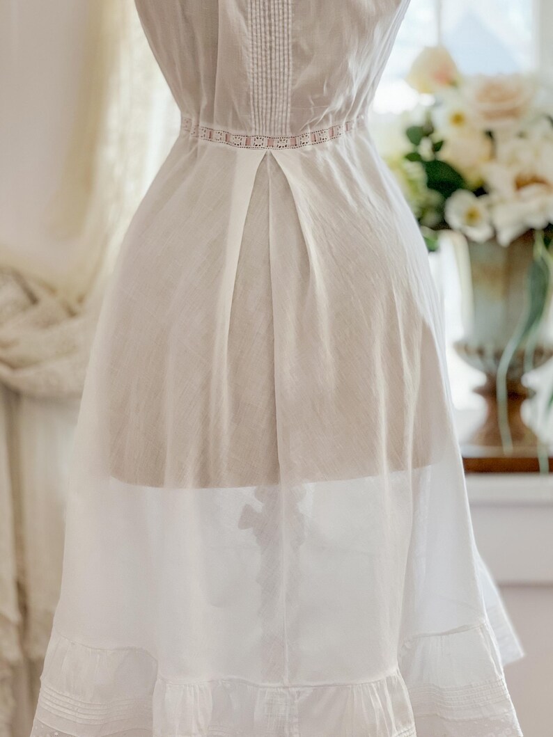 May include: A white vintage dress with delicate lace and pleating details. The dress features a high neckline with vertical stitching and a pink lace band at the waist. The skirt has a layered design with a ruffled hem, creating a flowing silhouette.