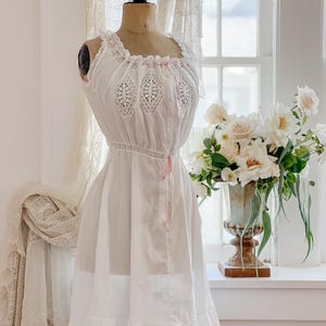 May include: A white vintage-style slip dress on a mannequin. The dress features lace detailing on the bodice and hem, with a delicate pink ribbon. The dress is displayed in front of a window with a vase of white flowers.