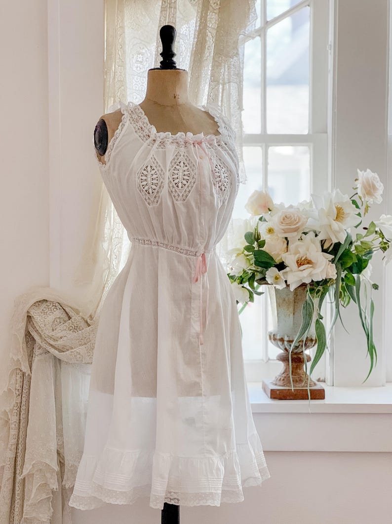May include: A white vintage-style slip dress on a mannequin. The dress features lace detailing on the bodice and hem, with a delicate pink ribbon. The dress is displayed in front of a window with a vase of white flowers.