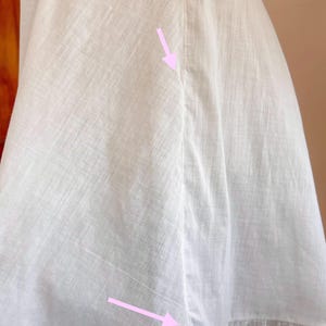 May include: Close-up of a white garment, possibly a dress or skirt, with visible seams and a ruffled hem. The fabric appears to be lightweight, with a subtle texture. The stitching details are prominent, highlighting the garment's construction.
