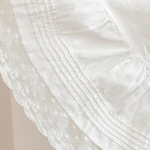 May include: Close-up of a white fabric with intricate details. The image showcases a layered design, featuring a delicate lace trim along the edge and a series of parallel pleats. The fabric appears soft and textured, suggesting a focus on textiles.
