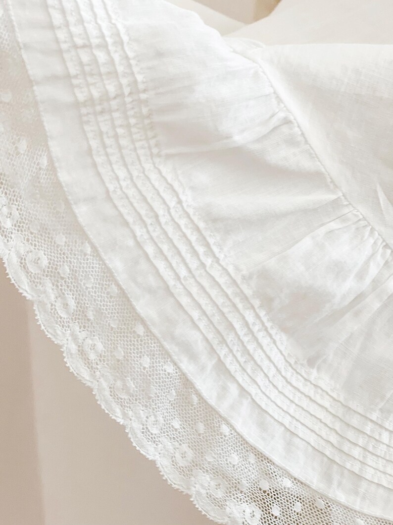 May include: Close-up of a white fabric with intricate details. The image showcases a layered design, featuring a delicate lace trim along the edge and a series of parallel pleats. The fabric appears soft and textured, suggesting a focus on textiles.