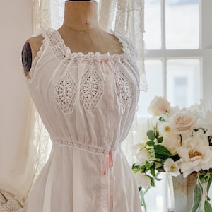 May include: White vintage-style nightgown on a mannequin. The nightgown features lace trim along the neckline and sleeves, with decorative lace panels on the bodice. Pink ribbon accents add a delicate touch. The dress has a gathered waist and a button-down front.