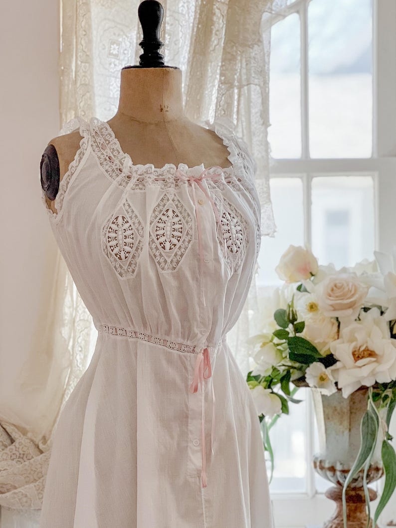 May include: White vintage-style nightgown on a mannequin. The nightgown features lace trim along the neckline and sleeves, with decorative lace panels on the bodice. Pink ribbon accents add a delicate touch. The dress has a gathered waist and a button-down front.