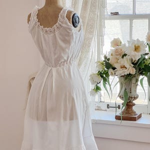 May include: A vintage white slip dress on a mannequin. The dress features delicate lace trim along the neckline, straps, and hem. The dress is displayed in front of a window with a bouquet of white flowers.