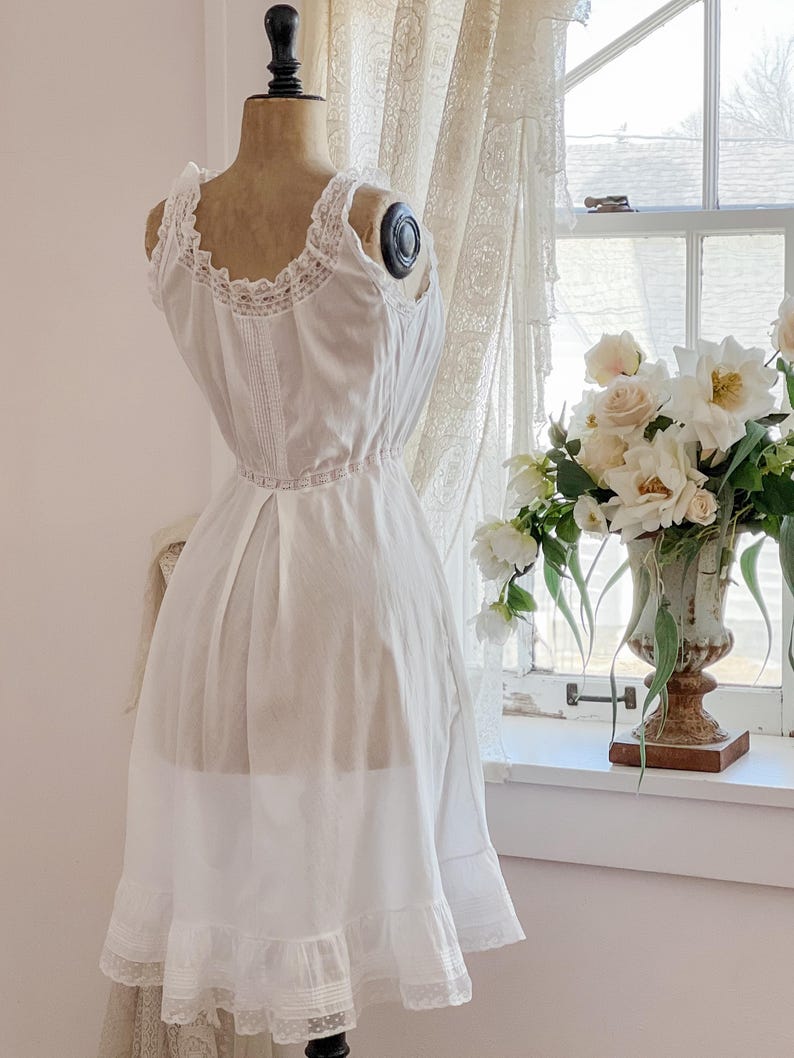 May include: A vintage white slip dress on a mannequin. The dress features delicate lace trim along the neckline, straps, and hem. The dress is displayed in front of a window with a bouquet of white flowers.