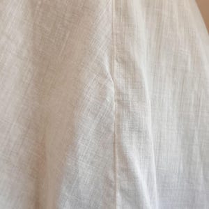 May include: Close-up of white linen fabric, showing the texture and weave of the material. The fabric is slightly wrinkled, with a vertical seam running down the center. The overall impression is of a soft, natural textile.
