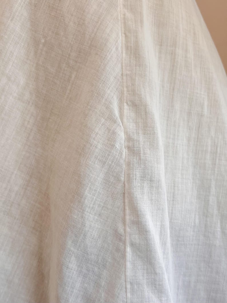 May include: Close-up of white linen fabric, showing the texture and weave of the material. The fabric is slightly wrinkled, with a vertical seam running down the center. The overall impression is of a soft, natural textile.