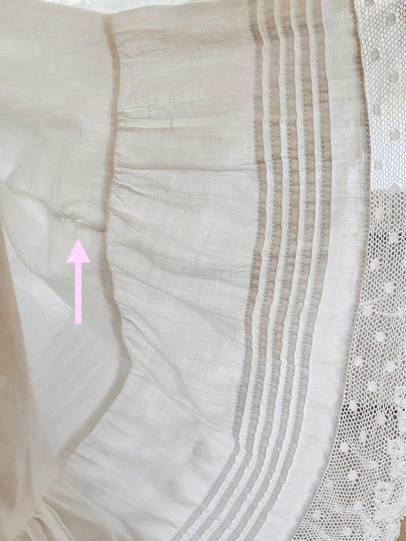 May include: Close-up of a white fabric with a ruffled edge. The fabric has vertical stripes and a delicate lace trim. The texture appears soft and lightweight, with subtle variations in tone.