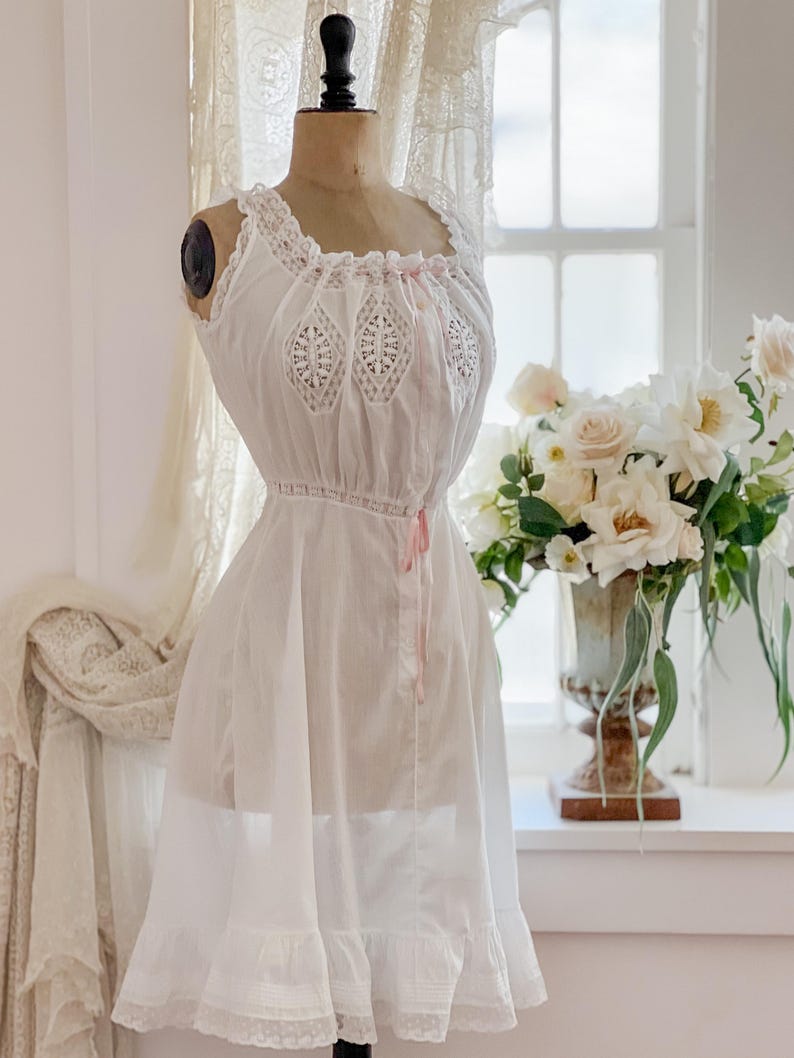 May include: A white vintage-style slip dress on a mannequin. The dress features lace detailing on the bodice and hem, with a delicate pink ribbon. The dress is displayed in front of a window with a bouquet of white flowers.