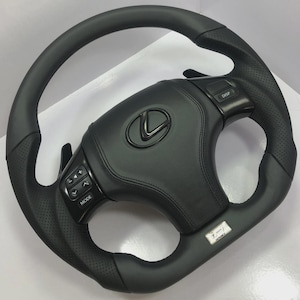 Puede incluir: Un volante Lexus de cuero negro con agarres laterales perforados, levas de cambio y botones de control integrados en los radios. El centro muestra un emblema Lexus negro y una insignia plateada F-Sport en la parte inferior.