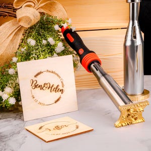 Puede incluir: Una caja de madera con un arreglo floral y un cuadrado de madera personalizado con el texto "Ben & Helen 08.12.2025". También hay un hierro de marcar de metal con un mango negro y naranja y una base dorada.