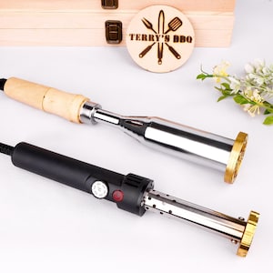 Puede incluir: Dos hierros para marcar para barbacoa. Uno es eléctrico con mango negro y control de temperatura. El otro tiene mango de madera y vástago metálico. Las cabezas para marcar son de latón. Una caja de madera y un círculo de madera con "TERRY'S BBQ" en el fondo.