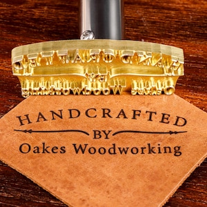 Może przedstawiać: Mosiężne żelazko do wypalania z napisem "HANDCRAFTED BY Oakes Woodworking" wytłoczonym na kawałku brązowej skóry. Żelazko do wypalania jest złote i ma metalowy uchwyt.