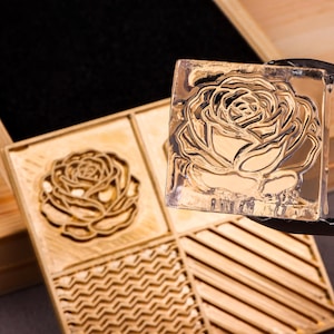 Könnte beinhalten: Ein goldfarbener, quadratischer Stempel mit detailliertem Rosenmuster wird in der Nähe einer Holzkiste mit geschnitzten Blumen- und geometrischen Mustern gehalten. Der Stempel dient wahrscheinlich zum Prägen.
