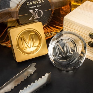 May include: A gold-coloured square ice stamp and a clear, round ice sphere with the letter "M" embossed. A bottle of cognac is in the background, along with a wooden box and metal tongs. The cognac bottle label reads "CAMVSE OLIVIA RAN XO COGNAC."