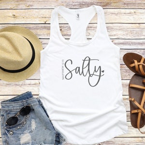 May include: White tank top with the text "Salty" in a cursive font and the text "Matthew 5:13" in a smaller font. The tank top is on a wooden background.