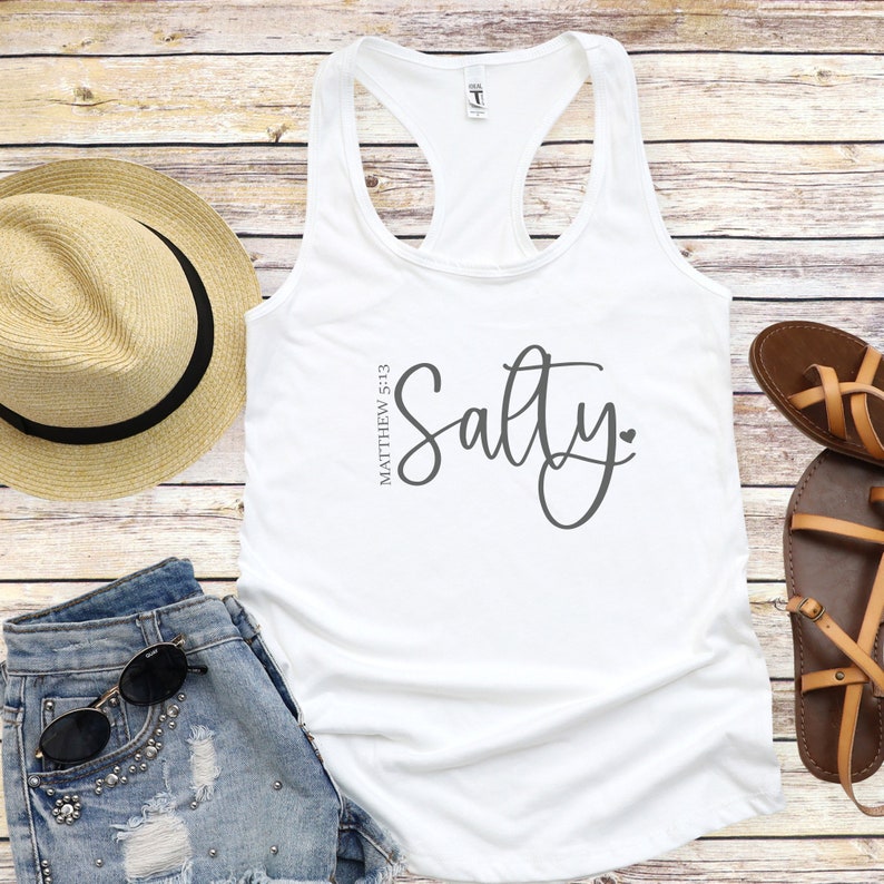 May include: White tank top with the text "Salty" in a cursive font and the text "Matthew 5:13" in a smaller font. The tank top is on a wooden background.