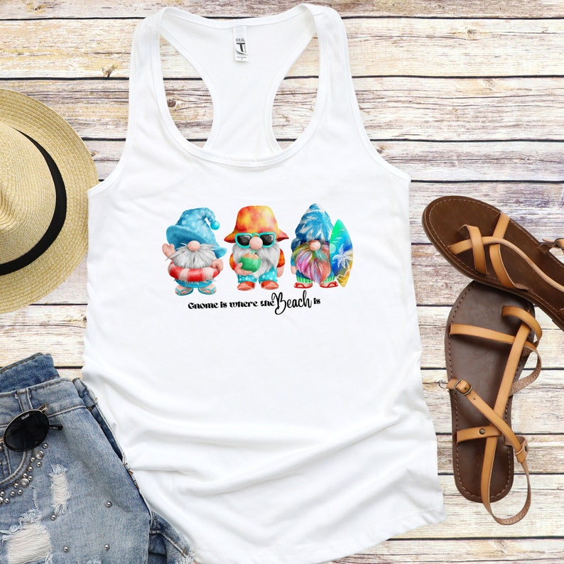 K&ouml;nnte beinhalten: Wei&szlig;es Tanktop mit einem Strandmotiv mit drei bunten Zwergen mit Strandaccessoires. Der Text "Gnome is where the Beach is" ist darunter gedruckt. Ein Strohhut, Jeansshorts, Sonnenbrille und Sandalen sind ebenfalls zu sehen.