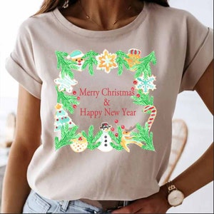 Op de afbeelding: Een beige T-shirt met een kerstontwerp met een krans van groene dennentakken, peperkoekjes en de tekst "Merry Christmas & Happy New Year".