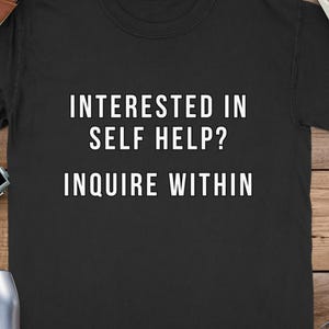 May include: Black t-shirt with the white text "INTERESTED IN SELF HELP? INQUIRE WITHIN". The shirt is on a wooden surface, with a camera, binoculars, a leather notebook, and a bag.