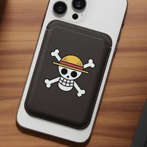 May include: A black phone wallet with a white skull and crossbones logo wearing a yellow straw hat, attached to a white smartphone. The wallet is rectangular and has a pocket for cards. The phone and wallet are on a wooden surface.