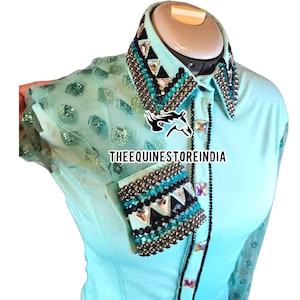May include: A turquoise equestrian show shirt with a black and turquoise crystal embellishment on the collar and cuff. The shirt has a sheer, sparkly overlay on the shoulders. The text "THEEQUINESTOREINDIA" is visible on the shirt.