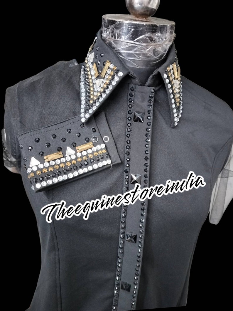 Pu&ograve; includere: Camicia nera senza maniche con colletto e polsini tempestati di strass. Il colletto presenta un design geometrico con accenti dorati. La camicia ha il testo "Thee equineststore india" stampato sul davanti.