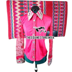 May include: A pink long-sleeved shirt with a lace overlay on the sleeves. The shirt has a rhinestone-studded collar and a row of rhinestones down the front. The shirt is on a mannequin and the text "THEEQUINESTOREINDIA" is visible on the shirt.