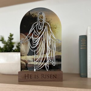 K&ouml;nnte beinhalten: Ein Holzst&auml;nder mit einer Glasscheibe, die eine wei&szlig;e Umrandung von Jesus zeigt, der aus einem Grab geht. Die Glasscheibe hat ein Hintergrundbild eines Grabes und einer Landschaft. Der Holzst&auml;nder tr&auml;gt die Worte "He is Risen" auf der Vorderseite.