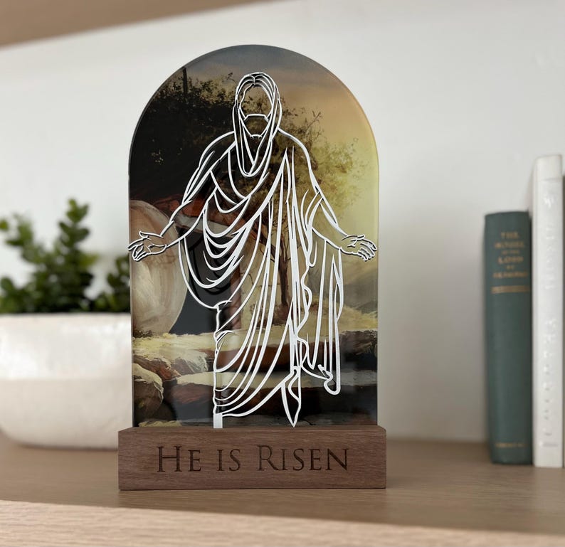 K&ouml;nnte beinhalten: Ein Holzst&auml;nder mit einer Glasscheibe, die eine wei&szlig;e Umrandung von Jesus zeigt, der aus einem Grab geht. Die Glasscheibe hat ein Hintergrundbild eines Grabes und einer Landschaft. Der Holzst&auml;nder tr&auml;gt die Worte "He is Risen" auf der Vorderseite.
