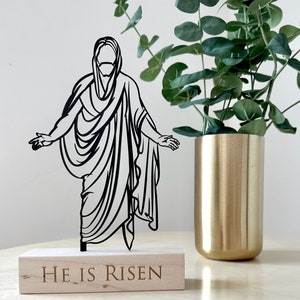 K&ouml;nnte beinhalten: Schwarze Silhouette von Jesus Christus, der auf einem Holzsockel mit den Worten "Er ist auferstanden" steht. Der Sockel steht auf einem hellen Holztisch mit einer goldenen Vase und einer gr&uuml;nen Pflanze im Hintergrund.