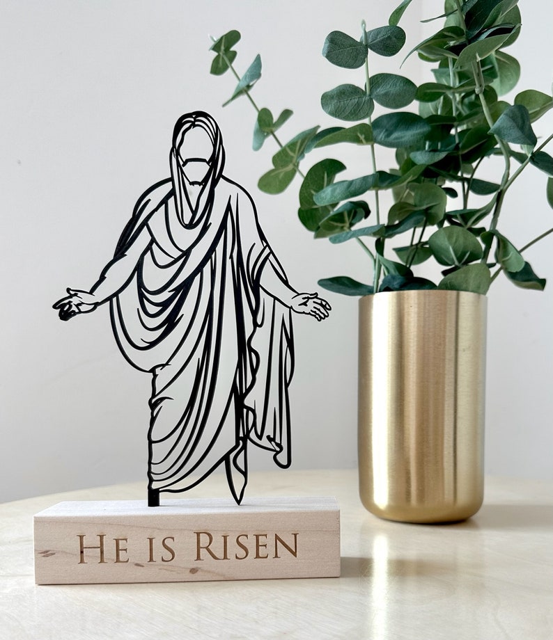 K&ouml;nnte beinhalten: Schwarze Silhouette von Jesus Christus, der auf einem Holzsockel mit den Worten "Er ist auferstanden" steht. Der Sockel steht auf einem hellen Holztisch mit einer goldenen Vase und einer gr&uuml;nen Pflanze im Hintergrund.