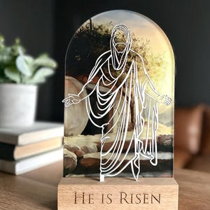 K&ouml;nnte beinhalten: Ein Holzst&auml;nder mit einer durchsichtigen Acrylplatte, die eine wei&szlig;e Umrandung von Jesus zeigt, der aus einem Grab geht. Die Platte hat ein Hintergrundbild eines Sonnenaufgangs und die Worte "He is Risen" sind in die Holzbasis graviert.