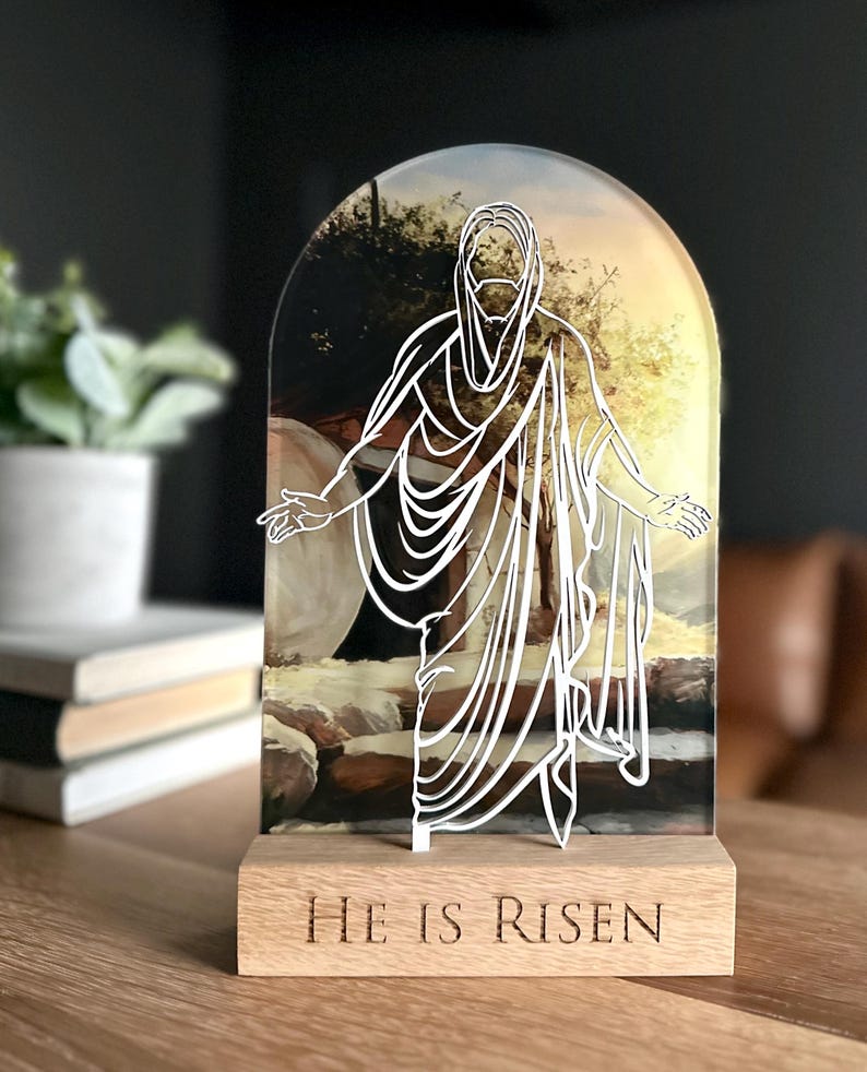 K&ouml;nnte beinhalten: Ein Holzst&auml;nder mit einer durchsichtigen Acrylplatte, die eine wei&szlig;e Umrandung von Jesus zeigt, der aus einem Grab geht. Die Platte hat ein Hintergrundbild eines Sonnenaufgangs und die Worte "He is Risen" sind in die Holzbasis graviert.