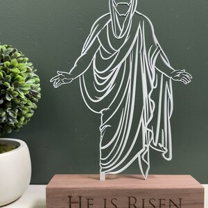 K&ouml;nnte beinhalten: Wei&szlig;e Silhouette von Jesus Christus, der auf einem Holzsockel steht, auf dem die Worte "Er ist auferstanden" eingraviert sind.