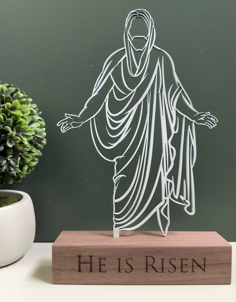 K&ouml;nnte beinhalten: Wei&szlig;e Silhouette von Jesus Christus, der auf einem Holzsockel steht, auf dem die Worte "Er ist auferstanden" eingraviert sind.