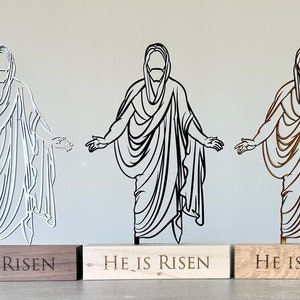 K&ouml;nnte beinhalten: Drei lasergeschnittene Holzfiguren von Jesus Christus mit ausgestreckten Armen. Jede Figur steht auf einem Holzsockel mit der Gravur "He is Risen". Die Figuren sind wei&szlig;, schwarz und braun.