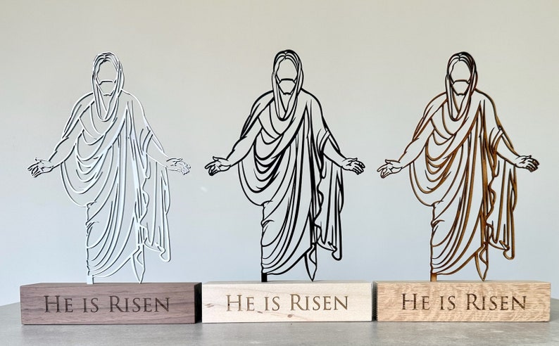 K&ouml;nnte beinhalten: Drei lasergeschnittene Holzfiguren von Jesus Christus mit ausgestreckten Armen. Jede Figur steht auf einem Holzsockel mit der Gravur "He is Risen". Die Figuren sind wei&szlig;, schwarz und braun.
