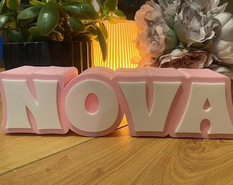 May include: Pink decorative letters spelling "NOVA" with white accents. The letters are displayed on a wooden surface, with plants and flowers in the background. The letters are a decorative item.