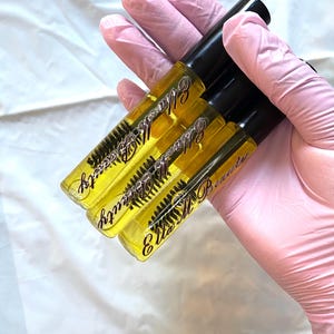 May include: Three clear tubes of eyelash serum with black caps are held in a gloved hand. The tubes contain a yellow liquid and a mascara wand. The text "Ella M Beauty" is printed on the tubes in a cursive font.