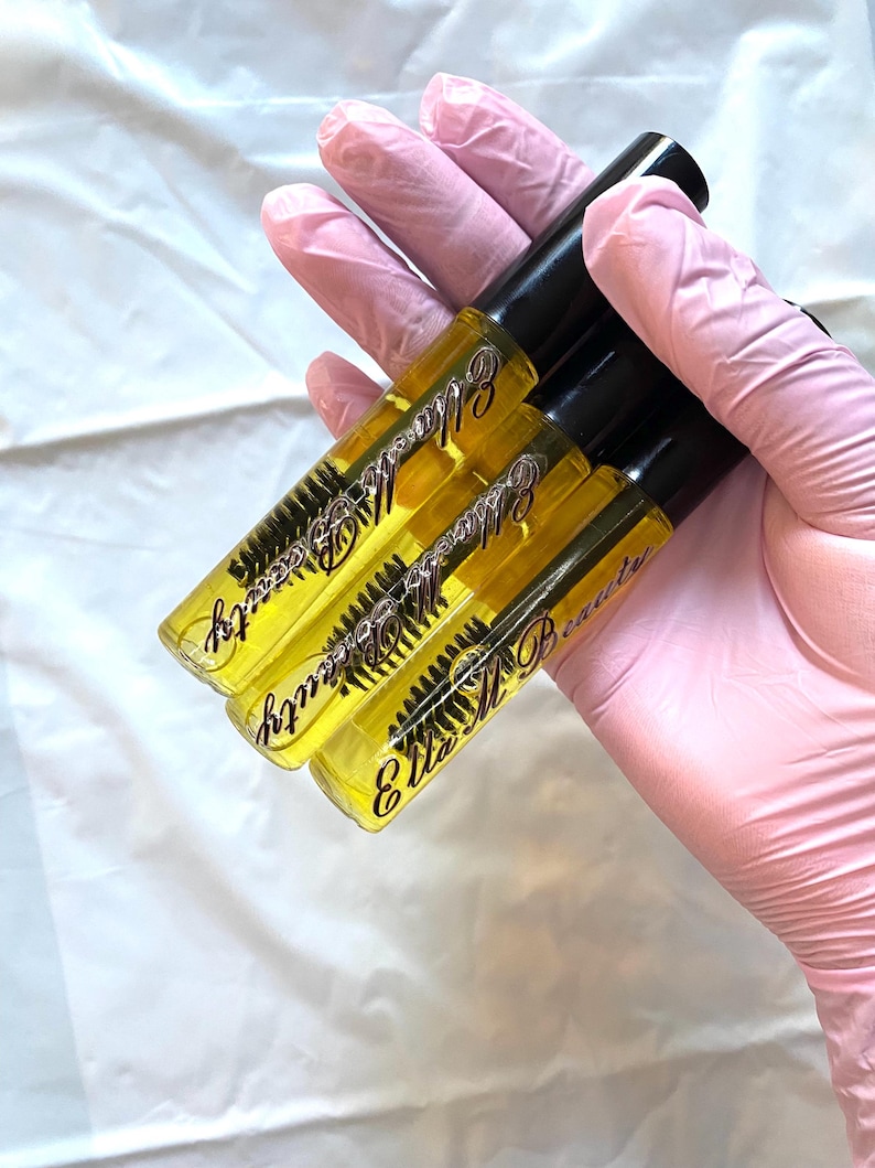 May include: Three clear tubes of eyelash serum with black caps are held in a gloved hand. The tubes contain a yellow liquid and a mascara wand. The text "Ella M Beauty" is printed on the tubes in a cursive font.