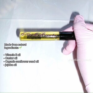 May include: A clear tube of eyelash and eyebrow oil with a black cap and a brush applicator. The tube contains a yellow liquid and has the text "Ella Ms. Beauty" printed on it. The text "Made from natural ingredients" is also visible.