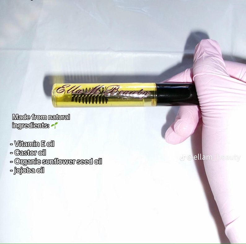 May include: A clear tube of eyelash and eyebrow oil with a black cap and a brush applicator. The tube contains a yellow liquid and has the text "Ella Ms. Beauty" printed on it. The text "Made from natural ingredients" is also visible.