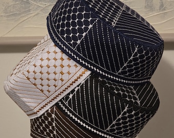 May include: Three embroidered skull caps in navy blue, white, and brown. The caps feature geometric patterns and decorative stitching. The caps are stacked on top of each other, showcasing the different designs and colors.