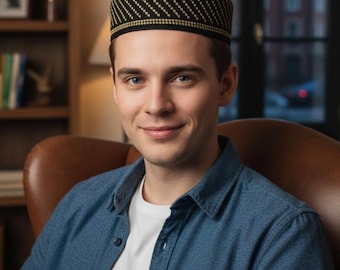 May include: A black and gold kufi hat with a geometric pattern. The hat is worn by a person wearing a blue denim shirt and a white t-shirt. The background includes a bookshelf and a window.